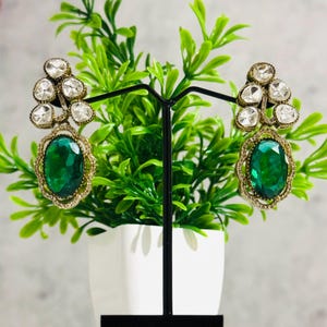Può includere: Un paio di orecchini pendenti con una grande pietra ovale verde e pietre più piccole trasparenti. Gli orecchini sono dorati e hanno un design delicato.
