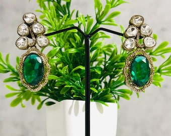 May include: A pair of dangle earrings with a large oval green gemstone and smaller clear stones. The earrings are gold-toned and have a delicate design.