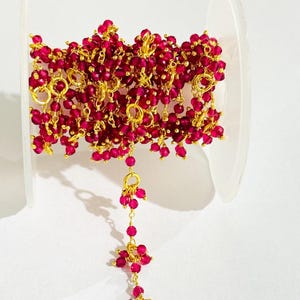 May include: A spool of gold chain with small, faceted, ruby red beads. The chain is strung with small, gold jump rings. The chain is hanging from a clear plastic spool.