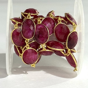 May include: A strand of faceted, oval-shaped deep magenta gemstones, each individually bezel-set in gold-toned metal. The stones are linked together in a continuous chain, showcasing a rich, saturated color and a reflective, textured surface.