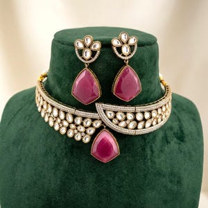 May include: A jewelry set consisting of a choker necklace and matching drop earrings. The pieces feature gold-tone metal, clear stones, and large, faceted, teardrop-shaped magenta stones. The necklace has a central magenta stone pendant.