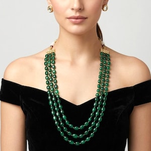 Puede incluir: Un collar de tres hileras con cuentas ovaladas de color verde esmeralda facetadas y detalles en tono dorado en el cierre. El conjunto incluye pendientes de botón redondos a juego con una piedra clara central y marco dorado.