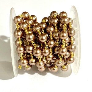 May include: A spool of brown faux pearl chain with gold-colored wire links. The pearls are a uniform size and color, and the chain is tightly wound on a clear plastic spool. Ideal for jewelry making and crafts.