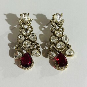 May include: A pair of gold tone dangle earrings with clear and red stones. The earrings have a teardrop shape and are adorned with multiple clear stones. The bottom of the earrings feature a large red teardrop stone.