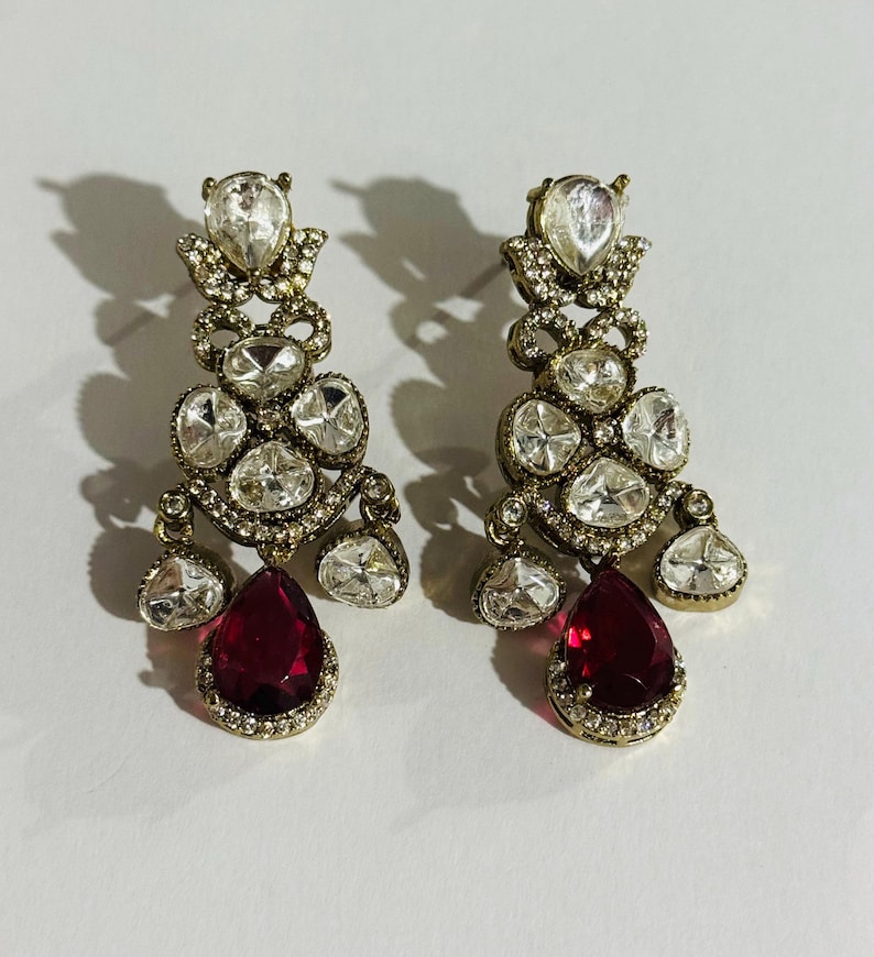 May include: A pair of gold tone dangle earrings with clear and red stones. The earrings have a teardrop shape and are adorned with multiple clear stones. The bottom of the earrings feature a large red teardrop stone.