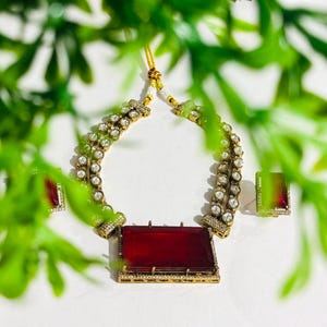 May include: A gold-coloured necklace and matching earrings set. The necklace features a large, rectangular, red gemstone pendant and two strands of small, clear gemstones. The earrings have a square red and green gemstone. The jewellery is set against a white background.