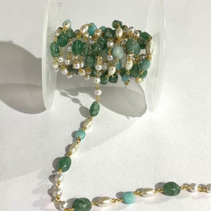 May include: A spool of beaded chain featuring green, white, and light blue gemstones and pearls. The chain is strung with gold-colored wire. The gemstones are various shapes and sizes, creating a unique and elegant design. Ideal for jewelry making.