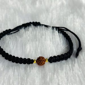 Könnte beinhalten: Schwarz geflochtenes Armband mit einer zentralen braunen Perle. Das Armband hat goldfarbene Akzente und verstellbare schwarze Kordeln. Dieses handgefertigte Armband ist ein Schmuckstück. Das Armband ist verstellbar.