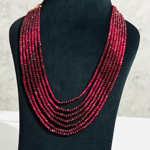 Può includere: Una collana a più fili composta da piccole perline sfaccettate di colore rosso intenso. La collana è esposta su un manichino nero, che mette in risalto il suo design a strati e il suo colore ricco. Le perline sembrano essere di dimensioni uniformi e ben infilate.