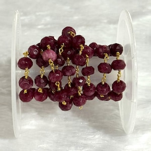 May include: A strand of faceted, deep magenta-colored gemstone beads linked together with thin gold-toned wire. The beaded chain is wrapped around a clear plastic spool, showcasing the uniform, round, and textured surfaces of each individual stone.