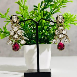 May include: A pair of dangle earrings with a gold tone setting and a teardrop shaped red stone. The earrings are adorned with clear rhinestones.