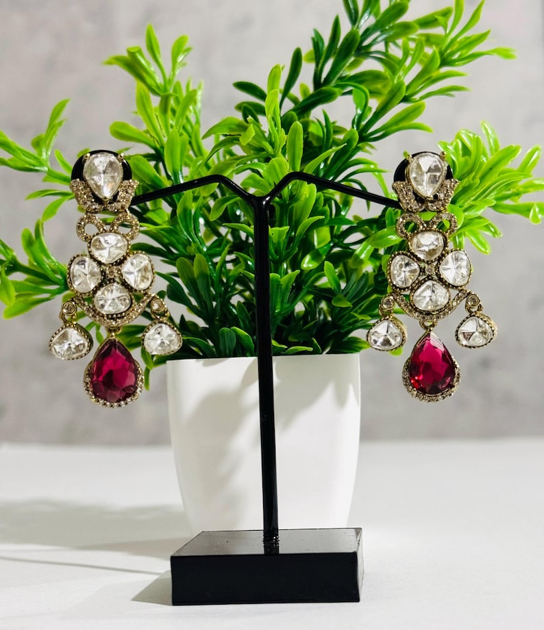 May include: A pair of dangle earrings with a gold tone setting and a teardrop shaped red stone. The earrings are adorned with clear rhinestones.