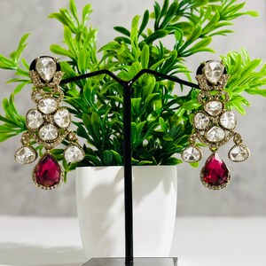 May include: A pair of dangle earrings with a teardrop-shaped red gemstone and a cluster of clear stones. The earrings are gold-toned and have a delicate design.