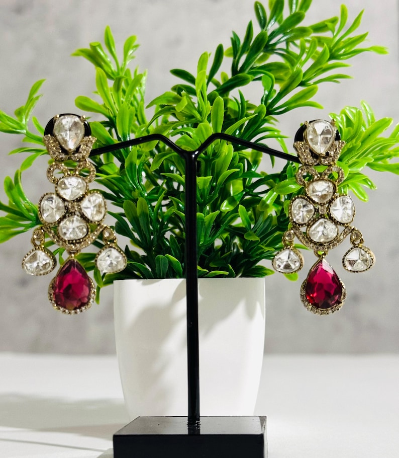 May include: A pair of dangle earrings with a teardrop-shaped red gemstone and a cluster of clear stones. The earrings are gold-toned and have a delicate design.