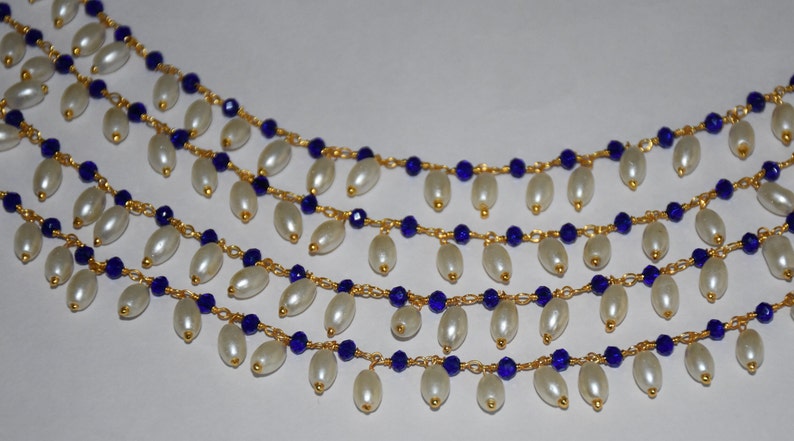 K&ouml;nnte beinhalten: Eine mehrreihige Halskette mit wei&szlig;en ovalen Perlen und blauen Glasperlen an einer goldenen Kette.
