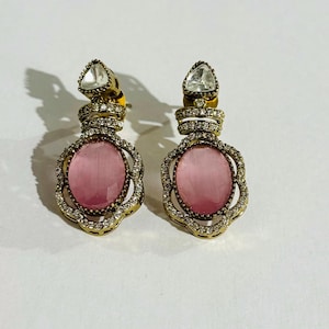 May include: A pair of gold-coloured drop earrings. Each earring features a pink, oval-shaped stone surrounded by small, clear crystals. A clear, triangular-shaped stone sits at the top of each earring. The earrings are designed with intricate detailing.