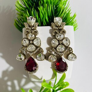 May include: A pair of dangle earrings with a gold tone setting and clear and red stones. The earrings feature a teardrop-shaped red stone at the bottom.