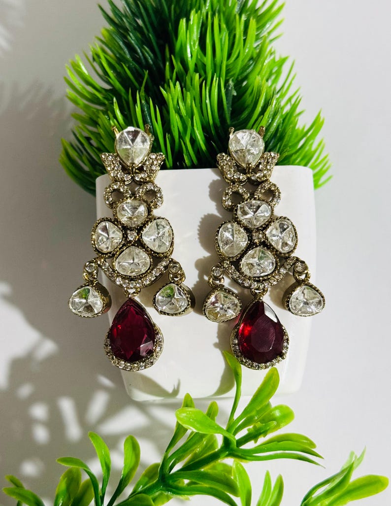 May include: A pair of dangle earrings with a gold tone setting and clear and red stones. The earrings feature a teardrop-shaped red stone at the bottom.