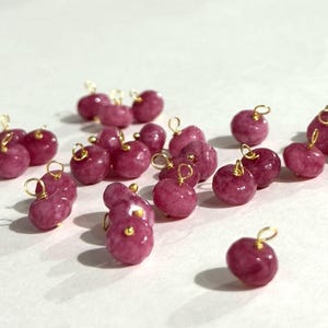 Puede incluir: Una colección de pequeñas cuentas redondas de piedra preciosa rosa con bucles de alambre dorados. Las cuentas están dispersas sobre una superficie blanca, mostrando su textura pulida y formas variadas. Adecuado para la fabricación de joyas.