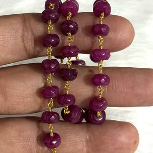 May include: A rosary-style chain featuring faceted, deep magenta-colored gemstone beads linked together with thin, gold-toned wire. The beads are round and slightly flattened, arranged in a continuous strand that loops back and forth.