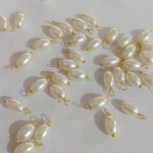 Puede incluir: Una colección de cuentas de perlas blancas ovaladas con bucles de metal dorado. Las cuentas están dispuestas sobre una superficie blanca.