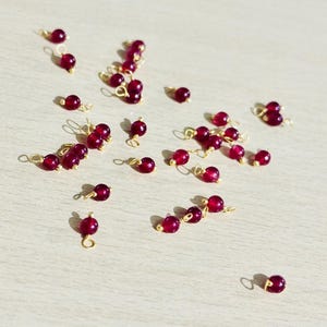 Op de afbeelding: Een verzameling van 25 rode edelsteen kralen met goudkleurige springringen. De kralen zijn ongeveer 6 mm in diameter en hebben een gladde, gepolijste afwerking. De springringen zijn bevestigd aan de bovenkant van elke kraal.