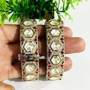 Puede incluir: Dos brazaletes dorados y plateados adornados con grandes piedras centrales hexagonales transparentes y piedras rojas más pequeñas. Los brazaletes están engastados con pequeñas piedras transparentes y tienen un cierre de bisagra. Fondo blanco brillante.