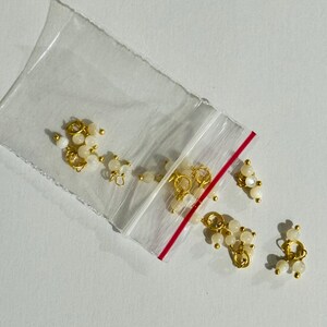 May include: A close-up of a bag of small gold and white bead charms. The charms are in the shape of a circle and have a small loop on the top. The beads are white and have a pearlescent finish.