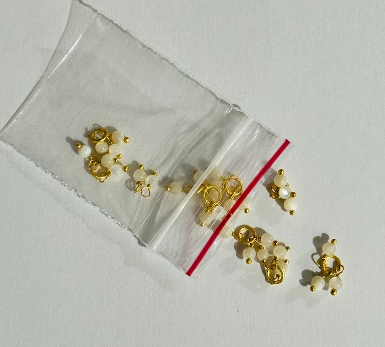 May include: A close-up of a bag of small gold and white bead charms. The charms are in the shape of a circle and have a small loop on the top. The beads are white and have a pearlescent finish.