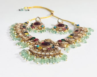 May include: An ornate gold-toned necklace and earring set. The necklace features a large central pendant with a deep red stone, surrounded by smaller colorful gems and light green teardrop-shaped accents. The earrings mirror the design.