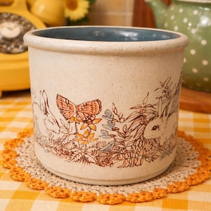 May include: A beige ceramic planter with a blue interior rim, decorated with a butterfly and rabbit illustration. It rests on a crocheted doily, placed on a yellow and white checked tablecloth. The planter is likely around 15 cm in height.