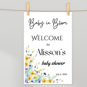 Puede incluir: Un letrero blanco de baby shower con el texto "Baby in Bloom" y "Welcome to Alisson's baby shower" en escritura elegante. Acentos florales de acuarela en amarillo, azul y morado adornan la esquina inferior izquierda. La fecha 5 de julio de 2025 está en la parte inferior.