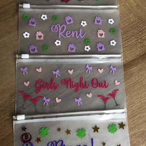 May include: Four translucent plastic zipper pouches with white zippers. The first pouch features the word Wages with piggy bank and coin icons. The second says Rent with house and flower icons. The third says Girls Night Out with martini glasses and bows. The fourth says Bingo! with bingo balls and stars.