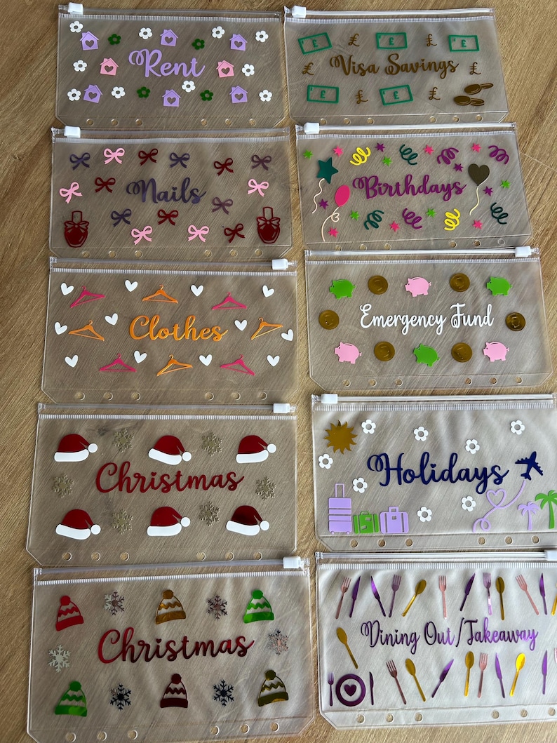 May include: Ten clear plastic zip-lock pouches for ring binders, each decorated with vinyl decals and text labels such as Rent, Visa Savings, Nails, Birthdays, Clothes, Emergency Fund, Christmas, Holidays, and Dining Out/Takeaway with varied icons.