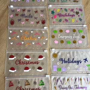 May include: A selection of clear, zipped pouches, each with a different label and decorative design. Labels include "Rent," "Nails," "Clothes," "Christmas," "Visa Savings," "Birthdays," "Emergency Fund," "Holidays," and "Dining Out/Takeaway."