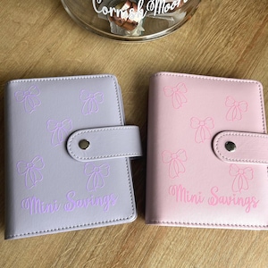 May include: Two small rectangular binders with snap closures, one in light purple and one in light pink. Both feature a repeating pattern of embossed bows and the text "Mini Savings" printed on the front cover in a matching colour.