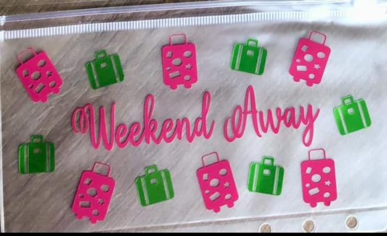 May include: A clear plastic zip-up pouch featuring the hot pink text "Weekend Away" in a cursive font, surrounded by a repeating pattern of hot pink and bright green suitcase icons with various circular and star-shaped cutouts on the luggage.