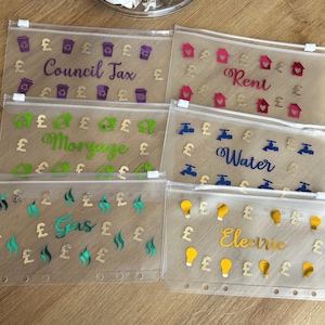 May include: Six clear plastic zippered pouches with gold pound signs and different coloured icons and text. The pouches are labelled "Council Tax", "Rent", "Mortgage", "Water", "Gas", and "Electric".