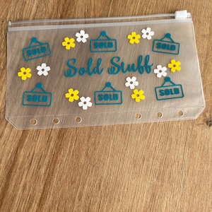 May include: A translucent plastic zip-up pouch featuring the text "Sold Stuff" in teal script, surrounded by teal "SOLD" signs and small yellow and white flower decals. The pouch has a white zip and six circular holes along the bottom edge.