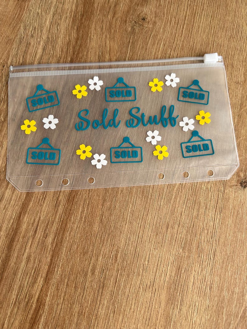 May include: A translucent plastic zip-up pouch featuring the text "Sold Stuff" in teal script, surrounded by teal "SOLD" signs and small yellow and white flower decals. The pouch has a white zip and six circular holes along the bottom edge.