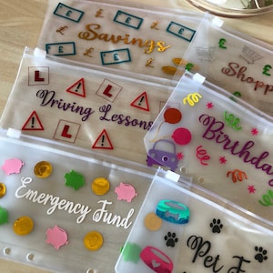 May include: A collection of translucent plastic zipper pouches for cash envelopes, each featuring colourful vinyl decals with text labels including Savings, Shopping, Driving Lessons, Birthdays, Emergency Fund, and Pet Fund, with various icons.