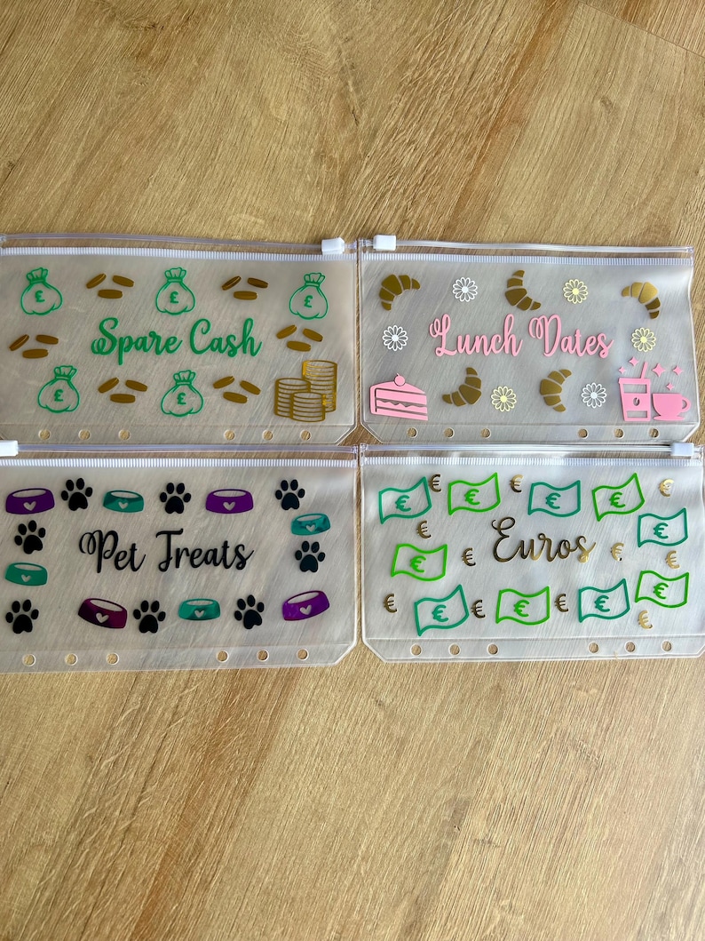 May include: Four rectangular, translucent plastic zipper pouches with white zippers and punched holes for binders. Each pouch features unique vinyl decals: green money bags and gold coins labelled Spare Cash, food items labelled Lunch Dates, paw prints and bowls labelled Pet Treats, and green euro bills labelled Euros.