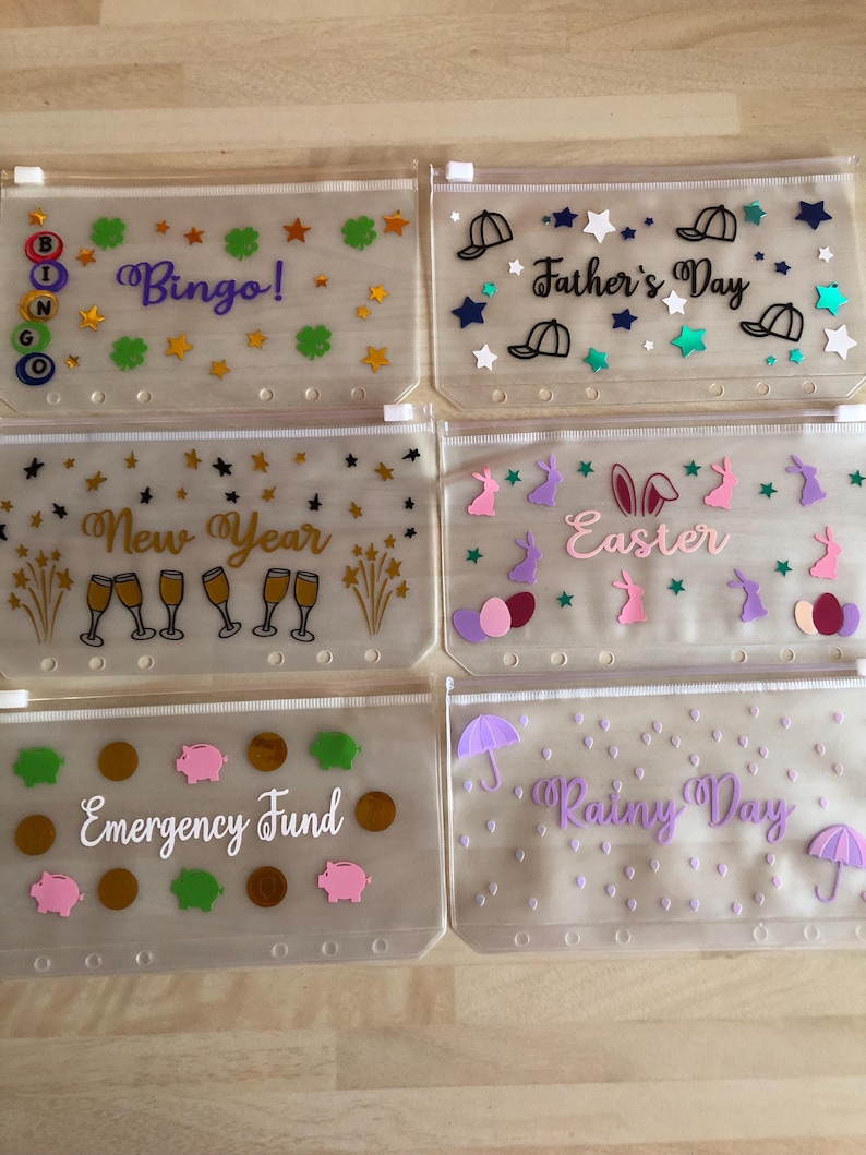 May include: Six translucent plastic zip-lock pouches with white plastic sliders and six holes along the bottom edge. Each pouch features unique vinyl decals: Bingo with BINGO letters, Father's Day with hats, New Year with champagne glasses, Easter with bunnies and eggs, Emergency Fund with coins and pigs, and Rainy Day with umbrellas and raindrops.