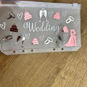 May include: A translucent plastic zip-up pouch featuring the word Wedding in white script. The pouch is decorated with pink and silver decals of tiered cakes, champagne flutes, interlocking wedding rings, hearts, and a silhouette of a couple.