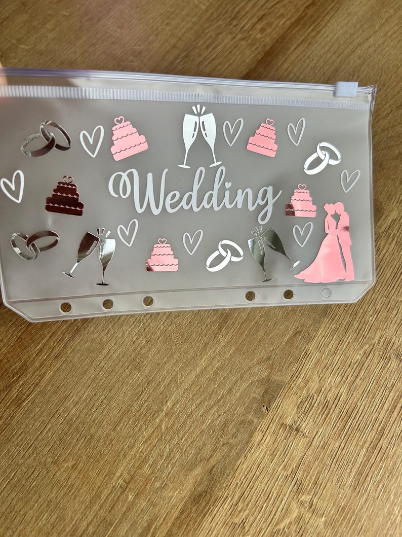May include: A translucent plastic zip-up pouch featuring the word Wedding in white script. The pouch is decorated with pink and silver decals of tiered cakes, champagne flutes, interlocking wedding rings, hearts, and a silhouette of a couple.