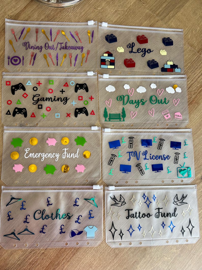 May include: Eight clear plastic zipper pouches for binders, each featuring unique labels and colourful vinyl decals. Labels include Dining Out/Takeaway, Lego, Gaming, Days Out, Emergency Fund, TV License, Clothes, and Tattoo Fund with matching icons.