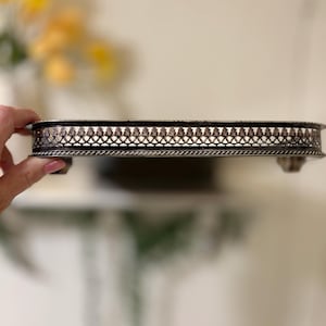 May include: A silver-toned, rectangular tray with ornate detailing. The tray features a decorative, openwork border and small feet. The design is vintage-inspired, suitable for serving or display. The tray is held by a hand.