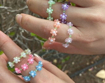 May include: Five colorful beaded rings with flower designs. The rings are made with small white and pastel colored beads. The rings are worn on the fingers of two hands.