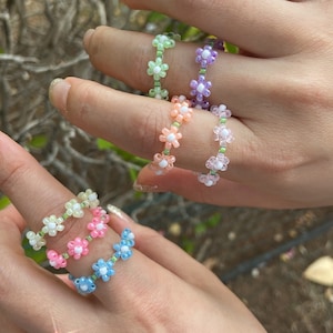 May include: Five colorful beaded rings with flower designs. The rings are made with small white and pastel colored beads. The rings are worn on the fingers of two hands.