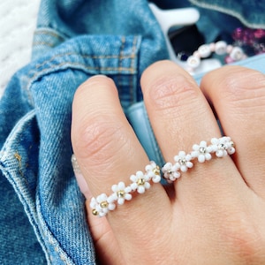 May include: Two white daisy flower beaded rings with gold accents. The rings are worn on a person's fingers.